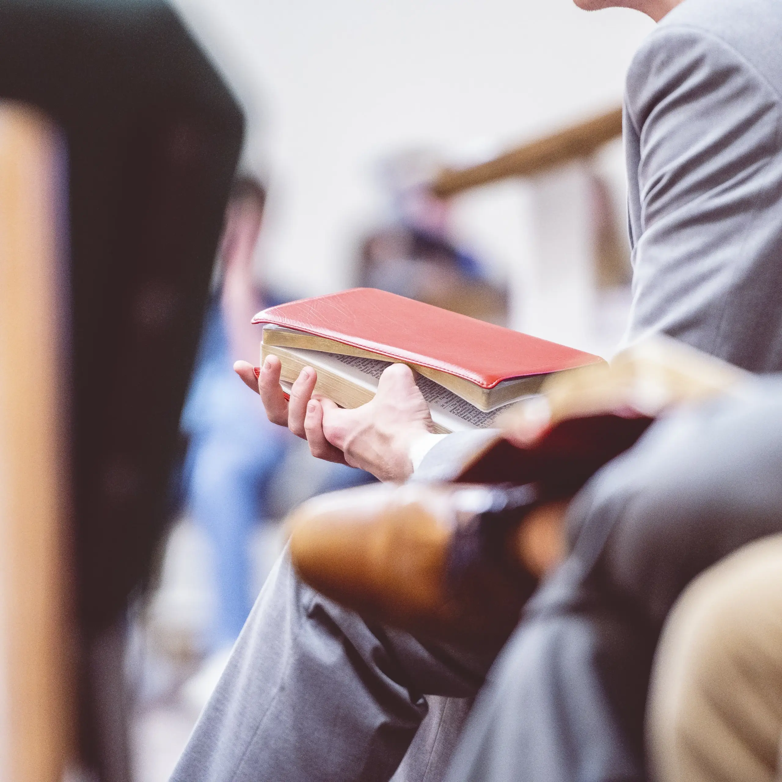A man's hands holding a Bible while sitting on a church pew with other people around him.