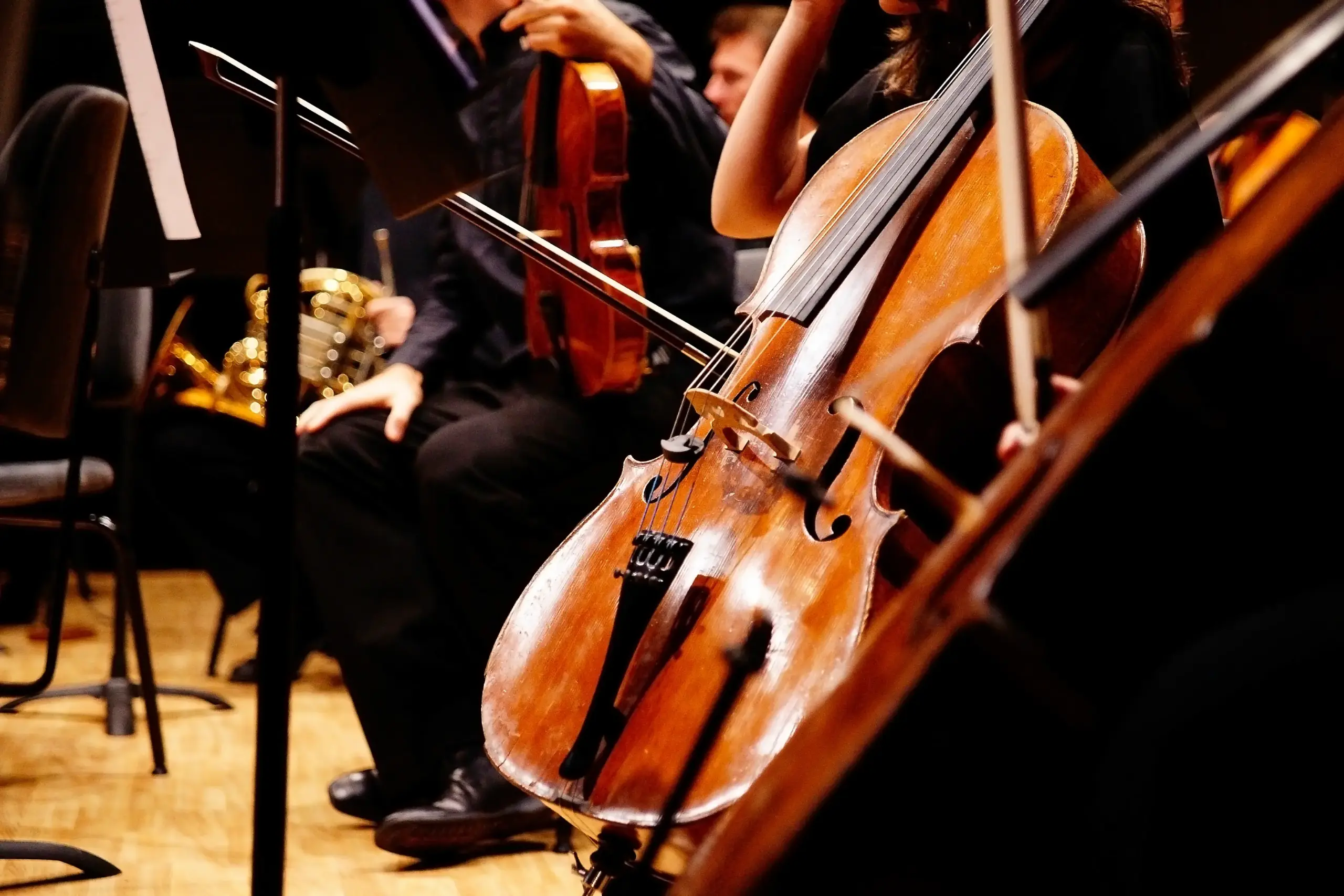 Close picture of a cello with someone sitting on a chair and holding a violin in the background.