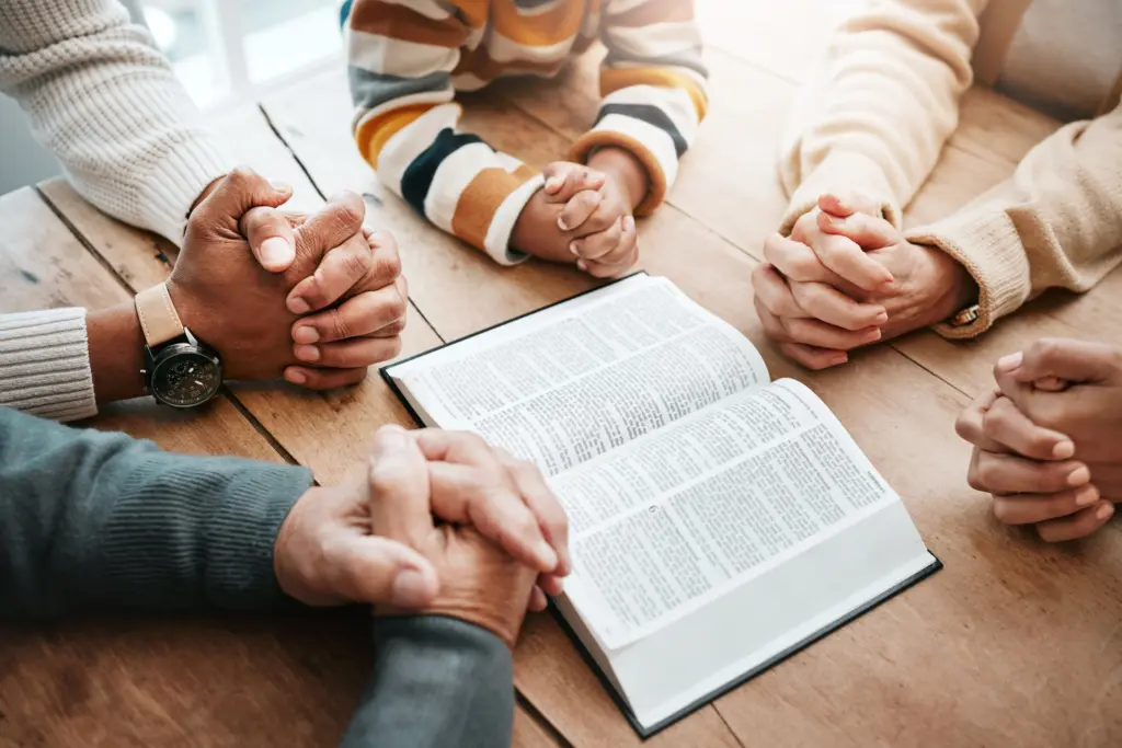 A table with an open Bible on it, and folded hands of people surrounding it and praying.
